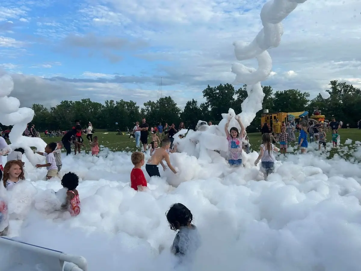Kids playing in a wide field of foam in a park, with a twisting column of foam rising behind them and families on the lawn.