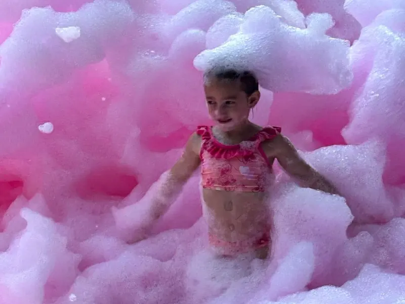 A young girl in a pink swimsuit standing in a wall of pink foam, with a heap of it on her head.