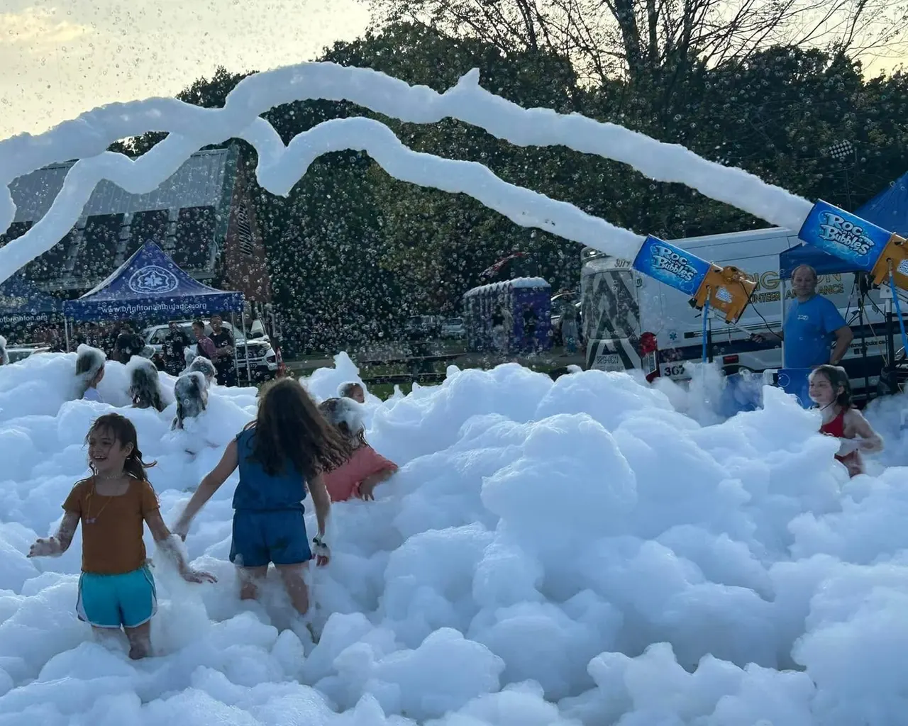 Kids wading through deep foam at dusk, with a man in a blue shirt standing by two ROC in the Bubbles cannons that send ropes of foam overhead.